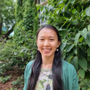 Profile headshot of CEBRA Research Fellow Lu-Yi Wang, a woman with long, black hair wearing a botanical print dress and green cardigan, standing in front of a vine-covered brick wall of a campus buildinhg