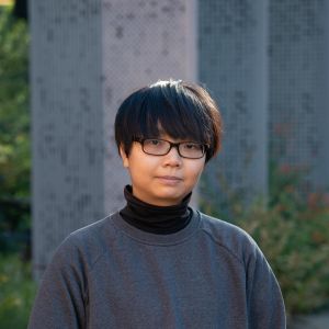 Profile head shot of CEBRA Research Fellow TK Le, a non-binary person with short, black hair wearing a grey sweatshirt over a black turtleneck against an out-of-focus background of three grey structures emerging out of green vegetation.